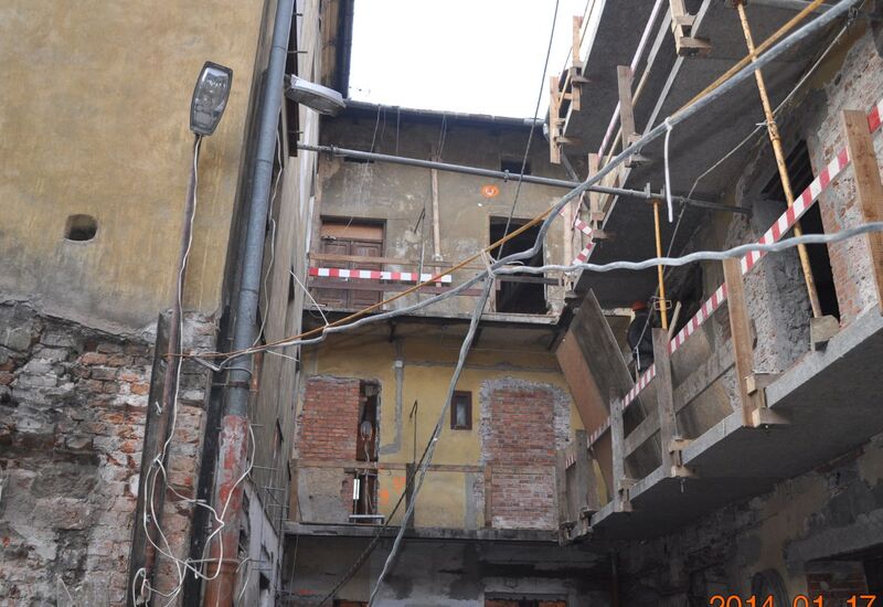 Dziedziniec starego budynku w remoncie, rusztowania, odsłonięte ceglane mury, taśmy przy balkonach i wiszące przewody.