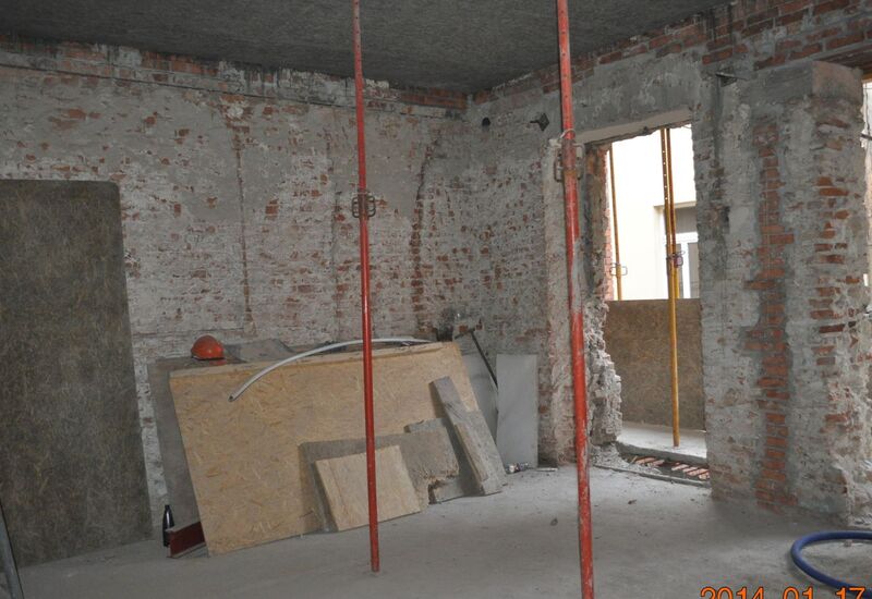 Wnętrze hotelu w remoncie z odsłoniętymi ceglanymi ścianami, czerwonymi podporami, zabitymi otworami okiennymi i składem materiałów budowlanych