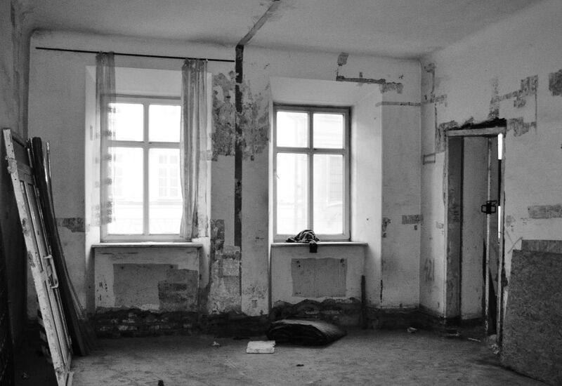 Puste pomieszczenie w remoncie z odpadającym tynkiem, dwoma oknami, drzwiami opartymi o ścianę, odsłoniętą cegłą i metalowym słupem