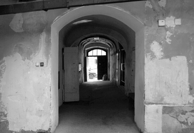 Łukowe przejście w starym budynku, z odpadającym tynkiem, ciemny korytarz otwiera się na jasną ulicę z zaparkowanym samochodem.