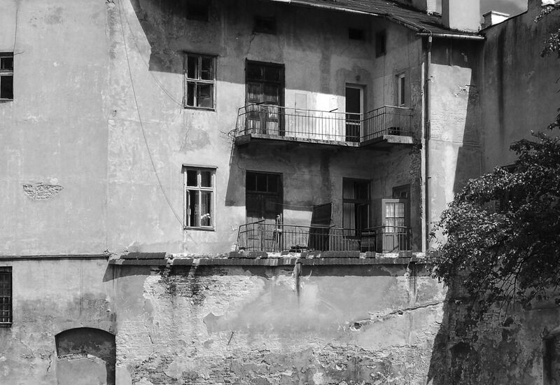 Zniszczona fasada kamienicy z dwoma balkonami, otwartymi drzwiami, odpadającym tynkiem, metalowymi balustradami i drzewem po prawej