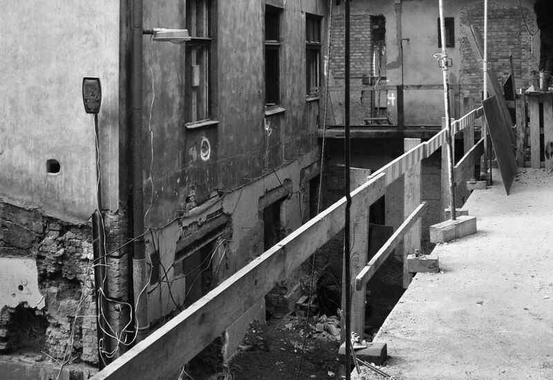 Wąska kładka rusztowania nad podwórzem starego hotelu w remoncie, odsłonięte ceglane ściany, poręcze i pracownik.