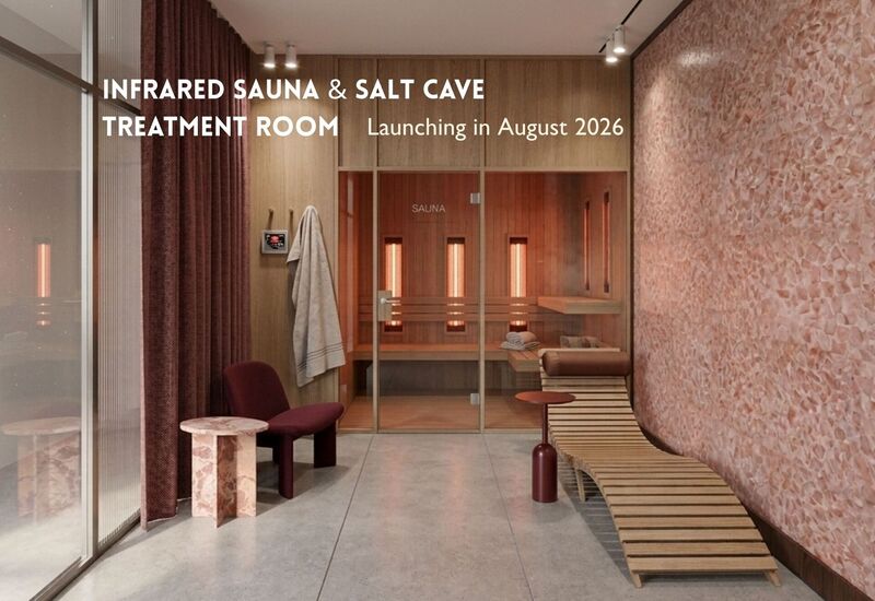 Przeszklona sauna na podczerwień z ciepłym światłem, różowa, solna ściana, drewniany leżak, bordowy fotel i marmurowy stolik.