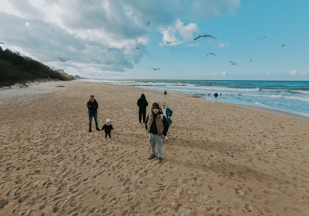 Sandy beachfront with guests strolling, choppy waves and seagulls overhead, dunes and a distant seaside building.
