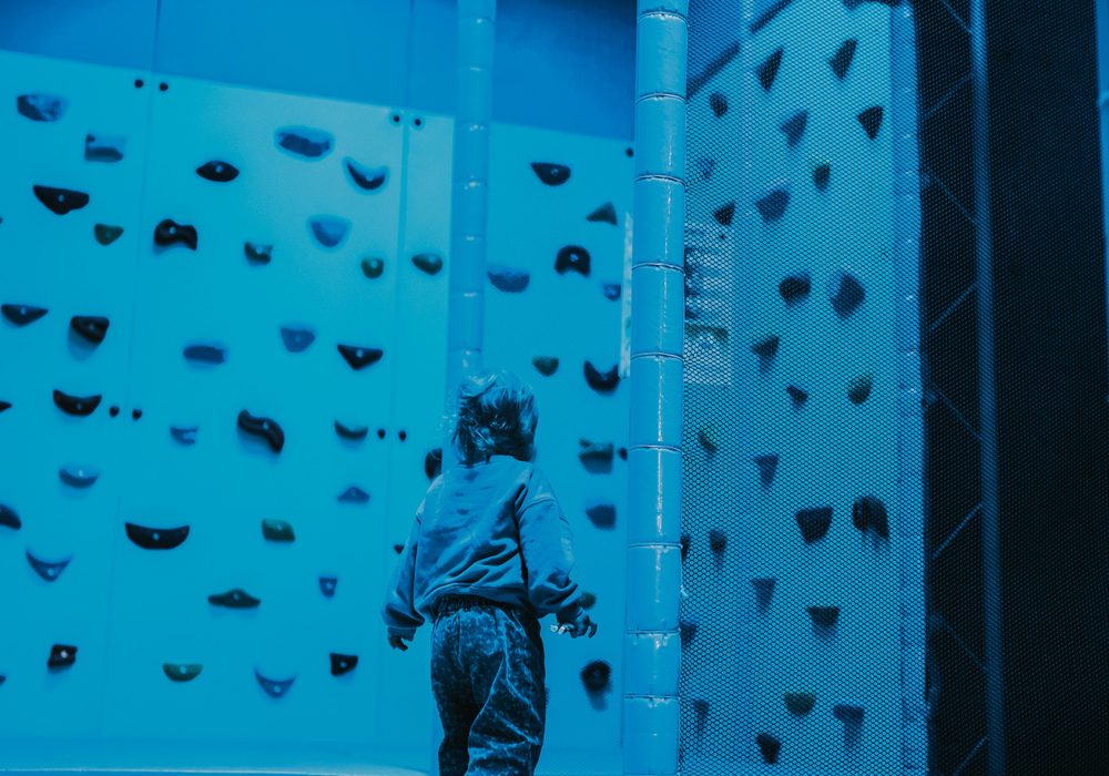 Family-friendly indoor play area with padded floors, climbing wall panels and netted play structure; a child explores under blue lighting