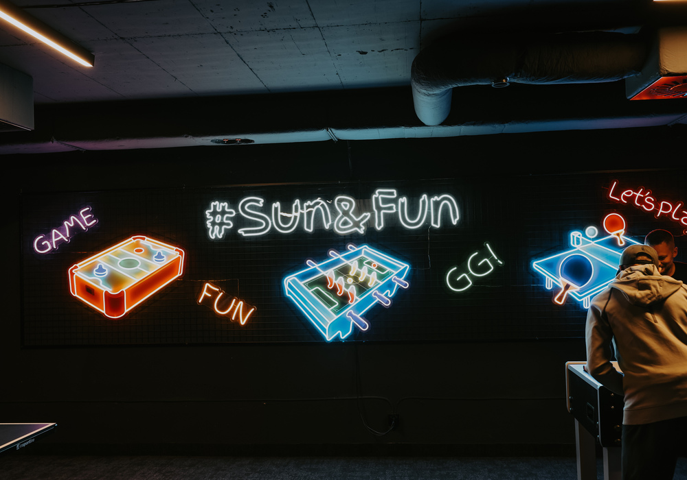 Hotel game lounge with neon '#Sun&Fun', colorful tabletop game icons and guests at a lit gaming table