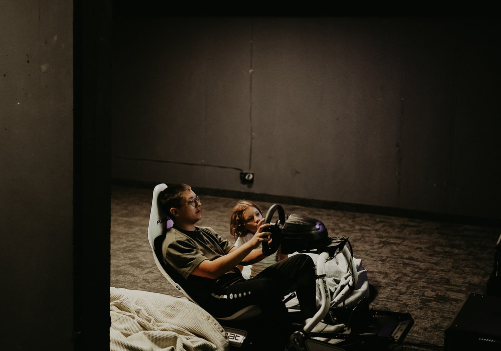 Dark hotel gaming lounge with exposed ceiling ducts, carpeted floor and two guests playing a racing simulator.