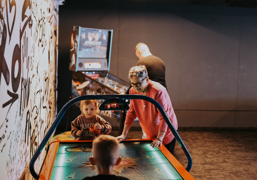Family playing air hockey in hotel game room with graffiti wall, pinball machine and warm overhead lighting