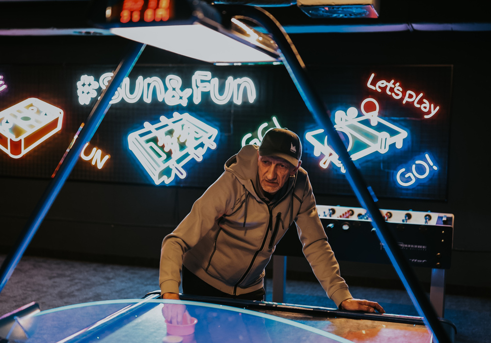 Man leaning over illuminated air hockey table in hotel game room with colorful neon signs and a foosball table