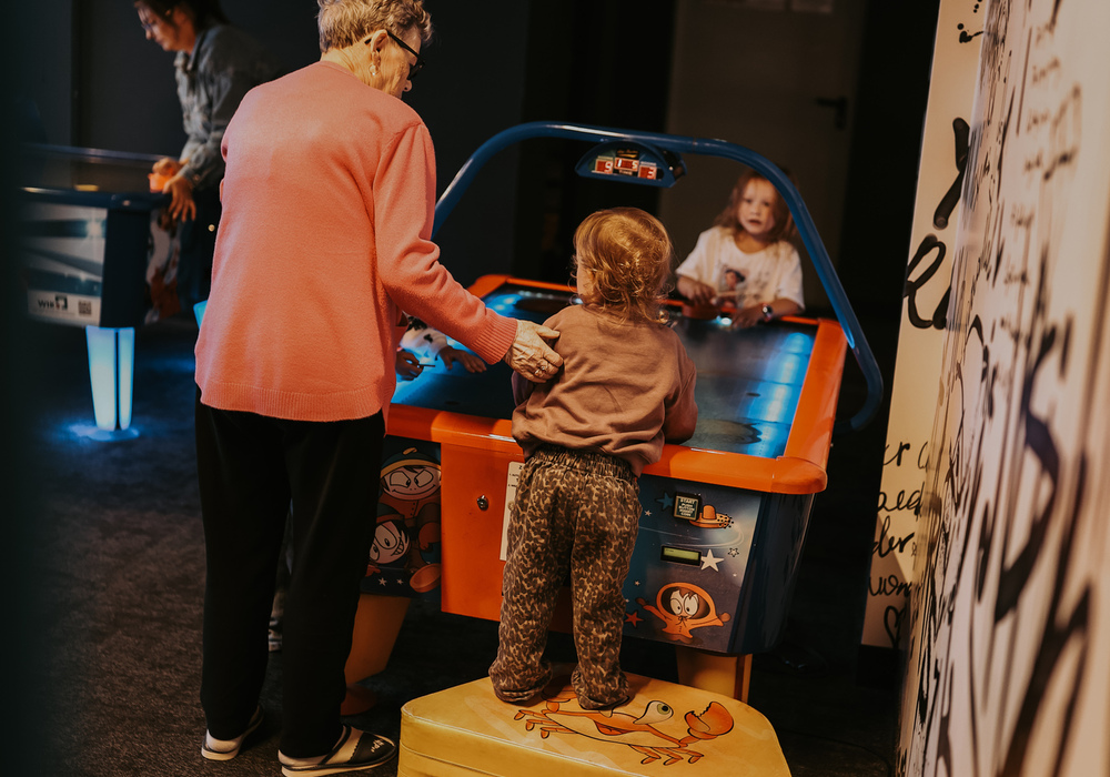 Grandmother helping a child play air hockey in a dim hotel family game room with arcade tables