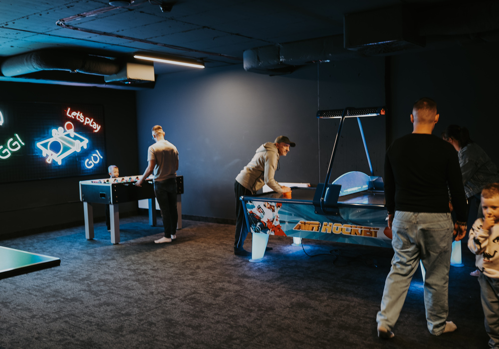 Hotel game room with neon 'Let's Play' sign, foosball and air hockey tables, relaxed family-friendly atmosphere