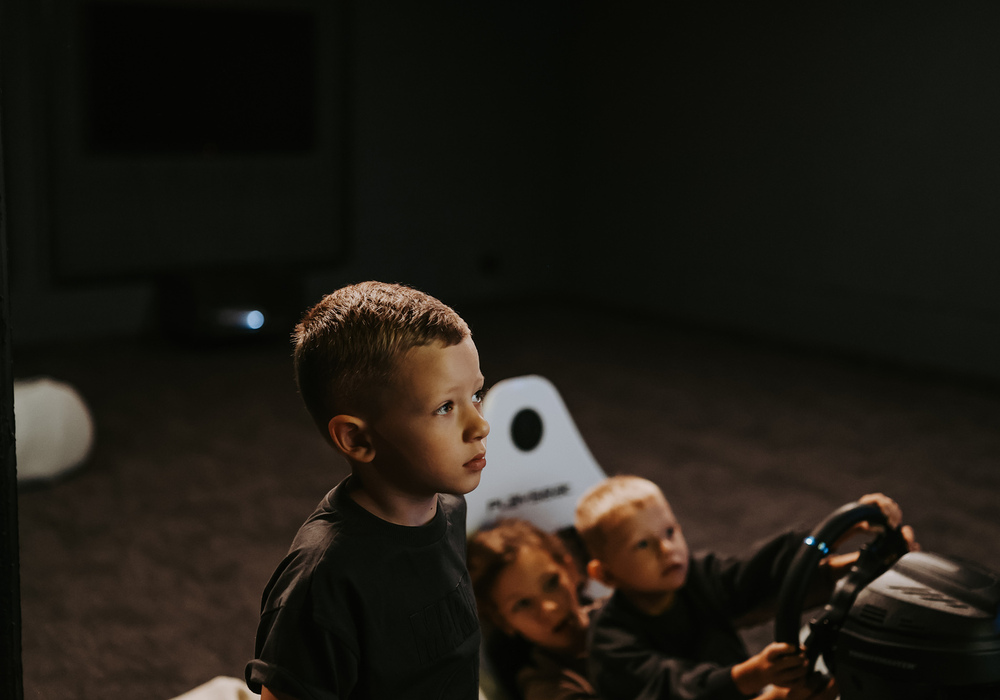 Children in a dim family entertainment room using a driving simulator beside beanbag seating and a neon sign