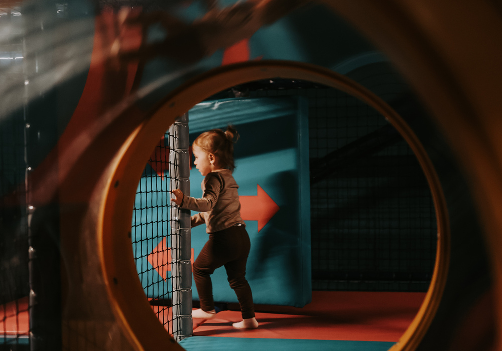 Toddler walking barefoot through a round tunnel toward padded climbing nets in a colorful indoor play area