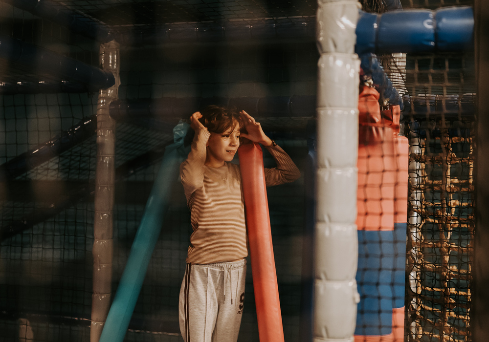 Child playing in a padded indoor soft-play area with netting, foam poles and hanging obstacles — family-friendly hotel playroom
