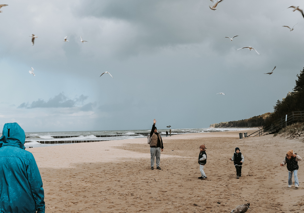 Sandy beach with seagulls swooping overhead, cloudy sky, an adult waving while children play near the shoreline