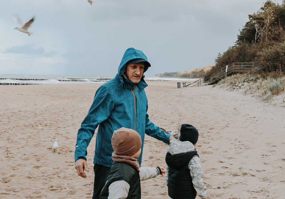 Grandfather and children walking a windswept beachfront, seagulls overhead, sandy shore and wooden dune walkway