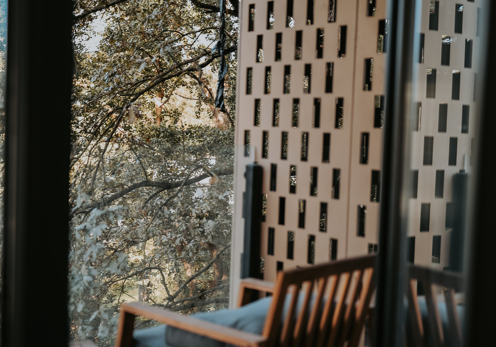 Private balcony with wooden lounge chair and cushions, perforated privacy screen and treetop view.
