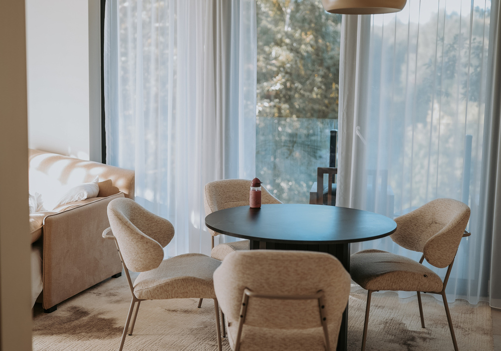 Cozy hotel dining nook with round table, four upholstered chairs, pendant lamp and sheer curtains opening to a balcony with tree views.