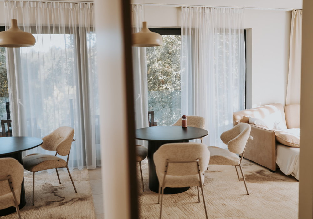 Cozy suite dining area with round black table, plush beige chairs, pendant lamps and sheer curtains to a tree-lined balcony