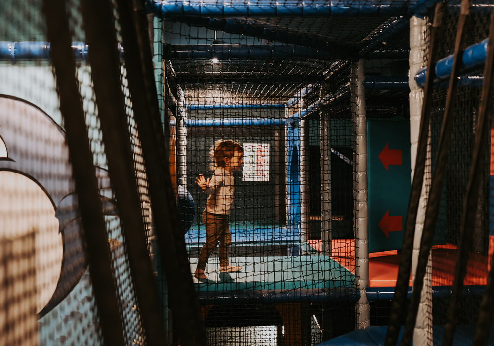 Indoor kids' play area with netted climbing tunnels, padded blue platforms and rope bridge where a child is playing.