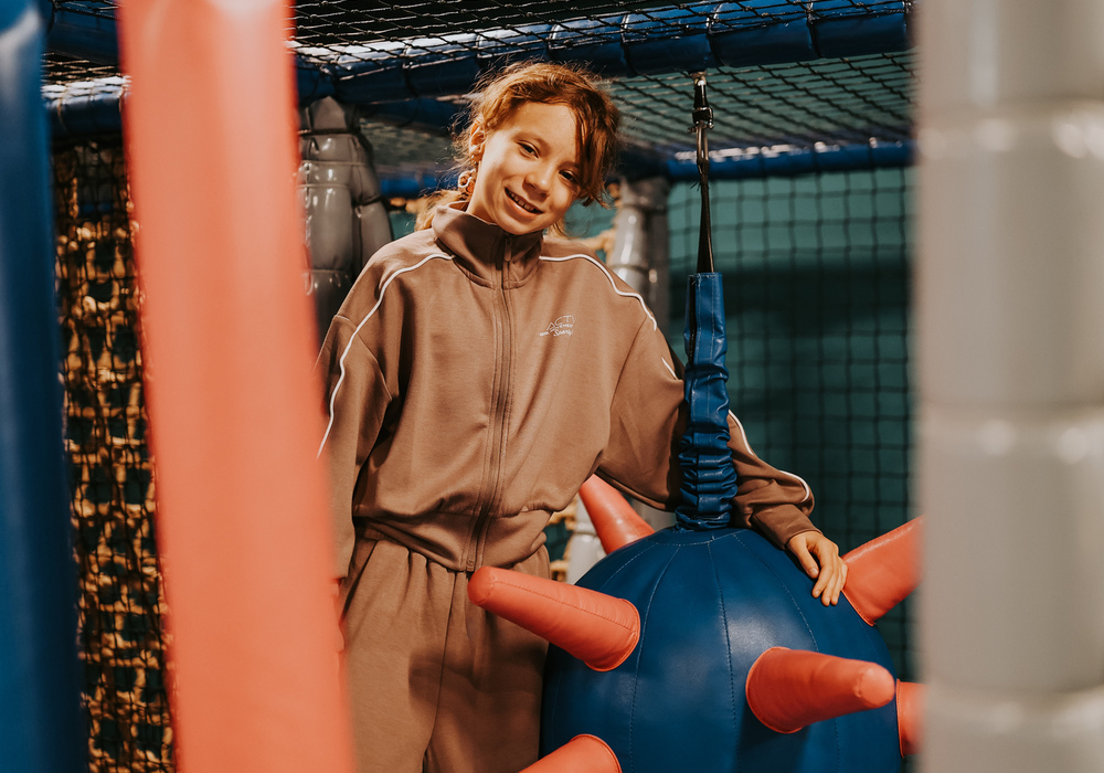 Smiling child playing in hotel's indoor soft-play area with netted climbing frame and blue inflatable with red soft spikes.