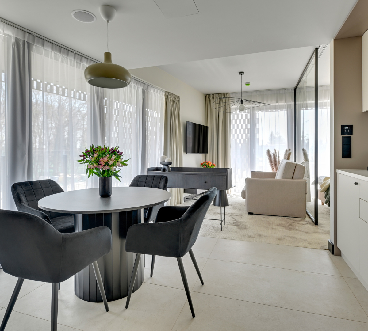 Bright modern suite with kitchenette, round table with flowers, black chairs, sofa, TV and floor-to-ceiling curtains.