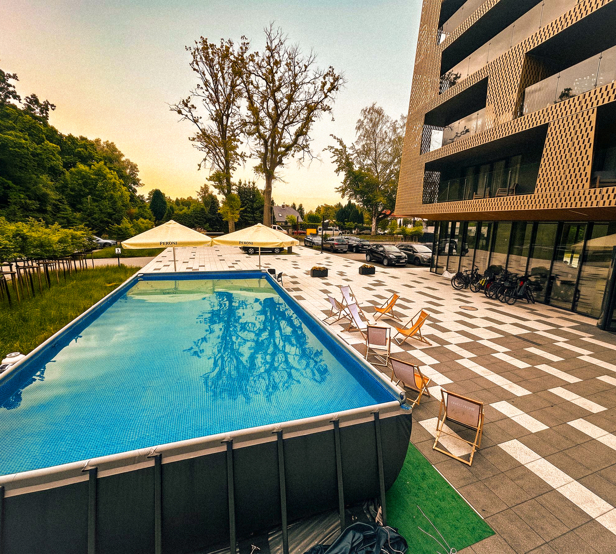 Nadziemny basen z leżakami i parasolami na tarasie obok nowoczesnej fasady hotelu i drzew