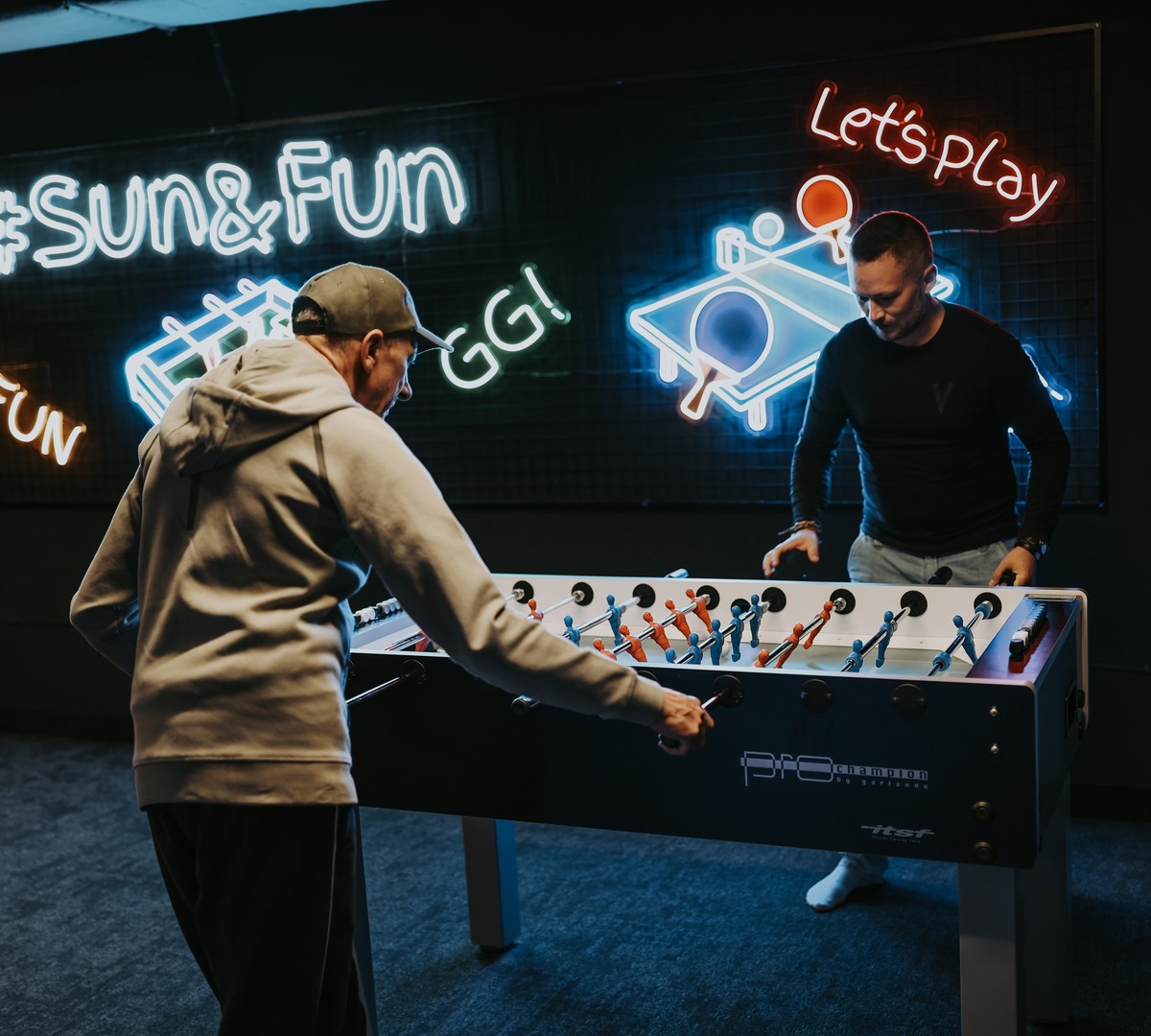 Guests playing foosball in neon-lit game room with colorful 'Let's play' and '#Sun&Fun' signs