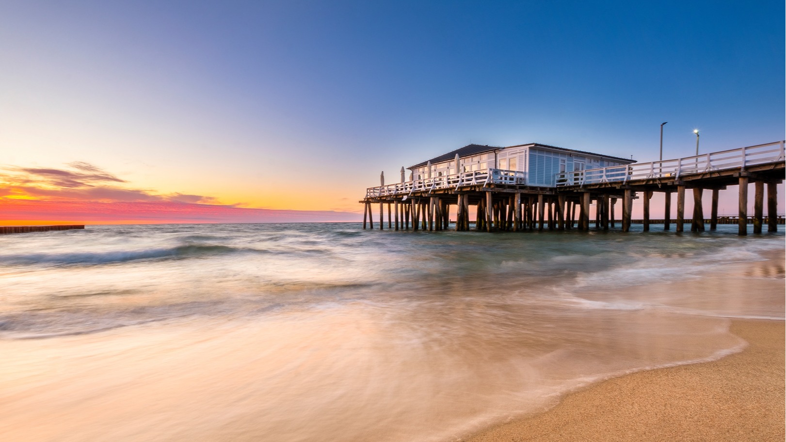 Molo na drewnianych palach, taras z parasolami, łagodne fale muskające piaszczystą plażę przy zachodzie słońca