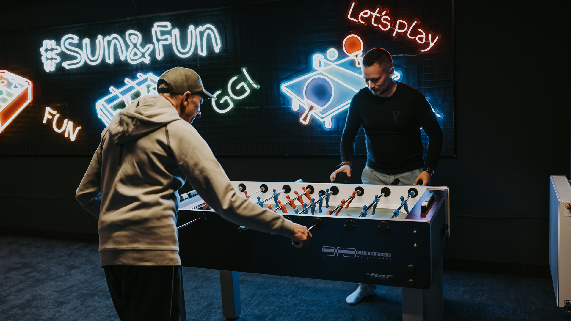 Guests play foosball in a hotel game room under neon 'Sun&Fun' and 'Let's play' signs, lively relaxed atmosphere