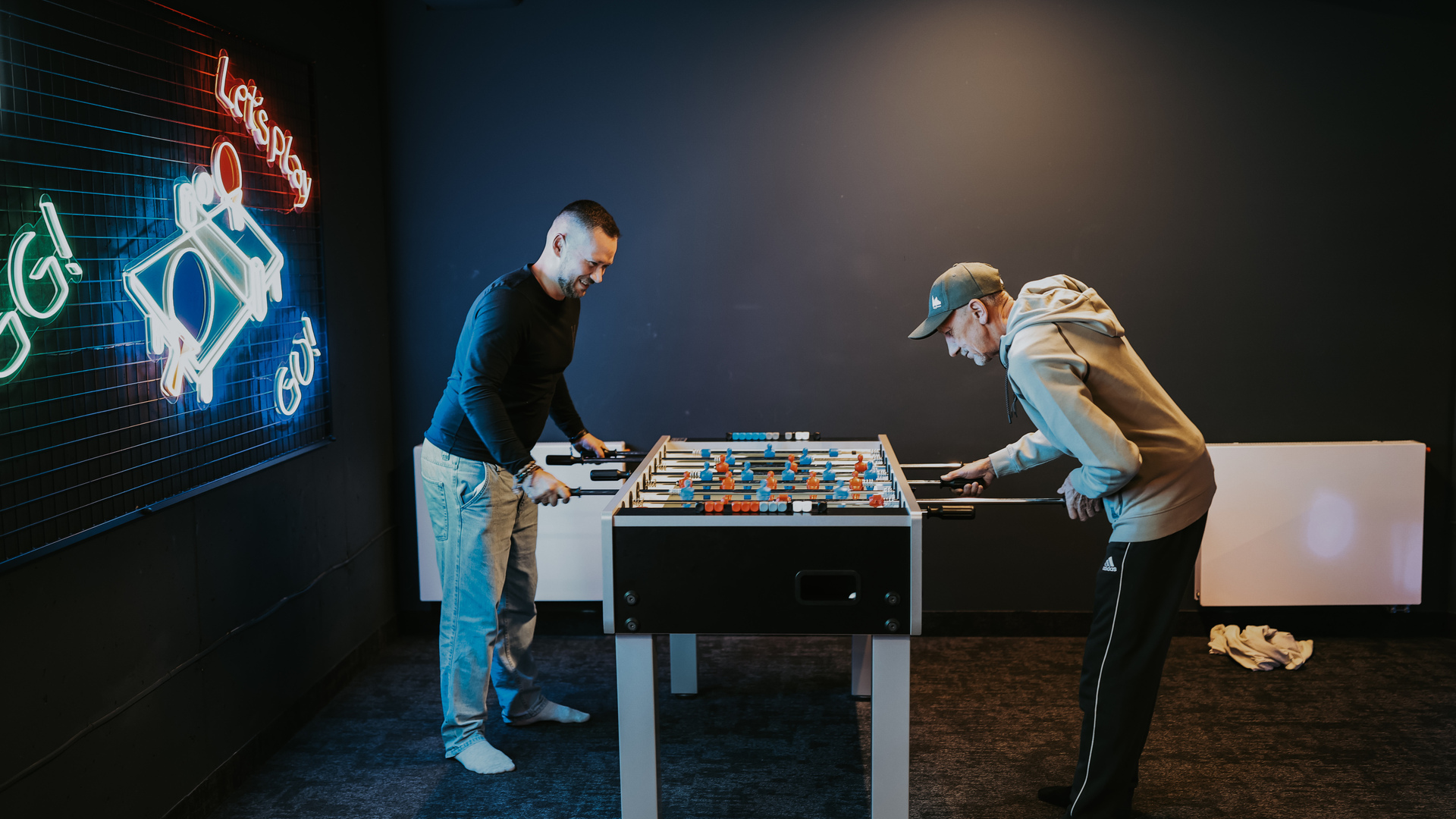 Casual game room with foosball table, neon 'Let's play' sign and two guests laughing while they play