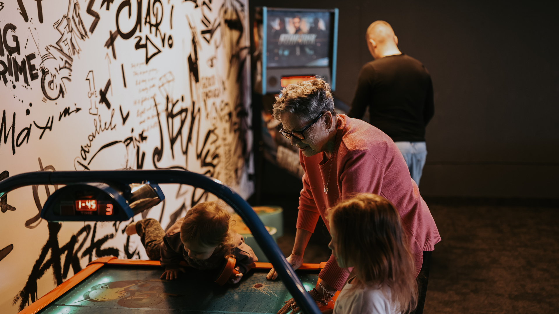 Game room with graffiti wall and arcade; grandmother helping kids at an air-hockey table in warm light.