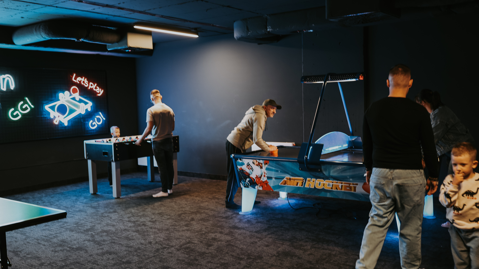 Hotel game room with air hockey and foosball, neon 'Let's Play' sign and guests enjoying casual play