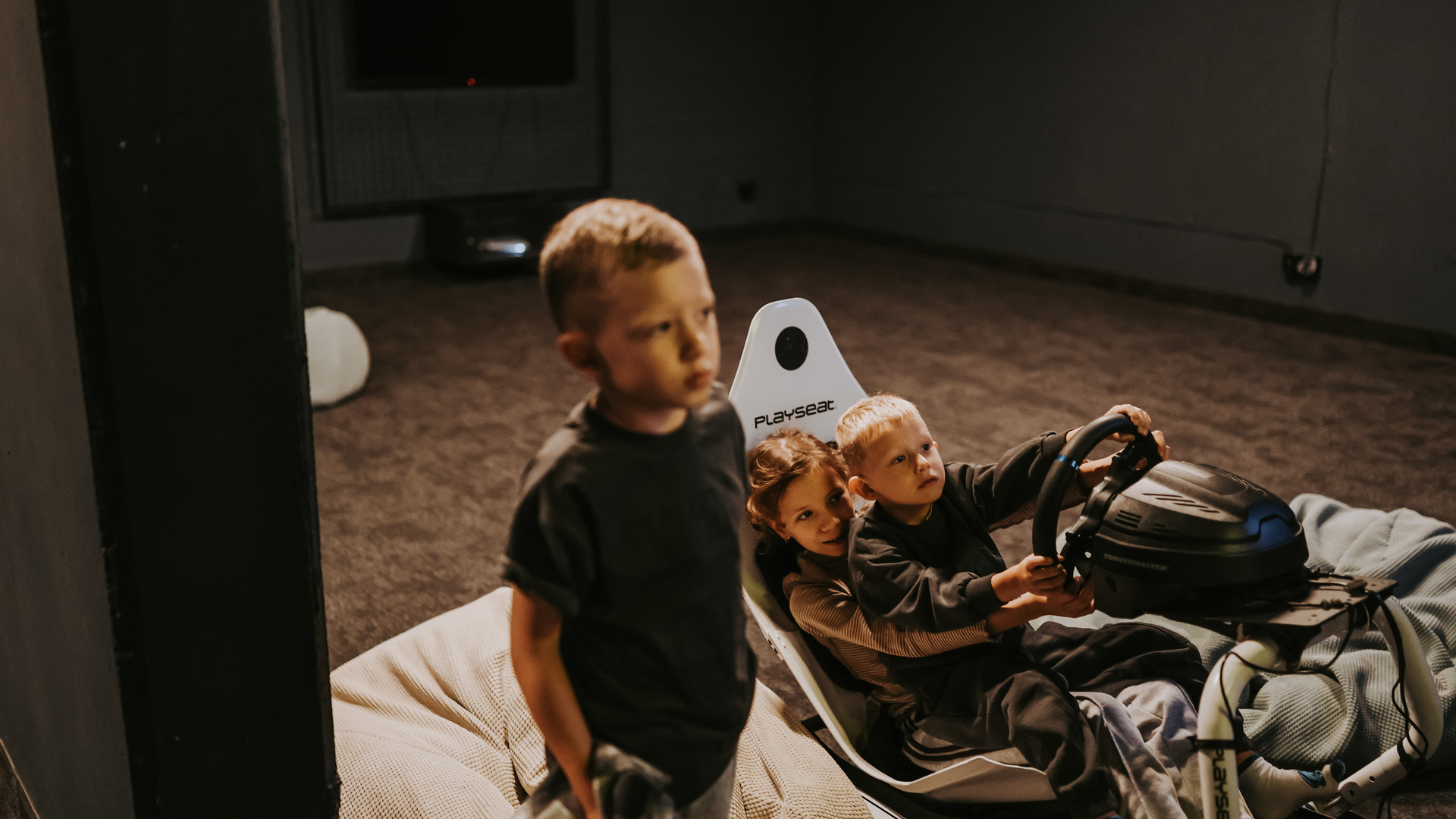 Children sharing a Playseat racing simulator in a dim family game room with beanbag seating and soft carpet.