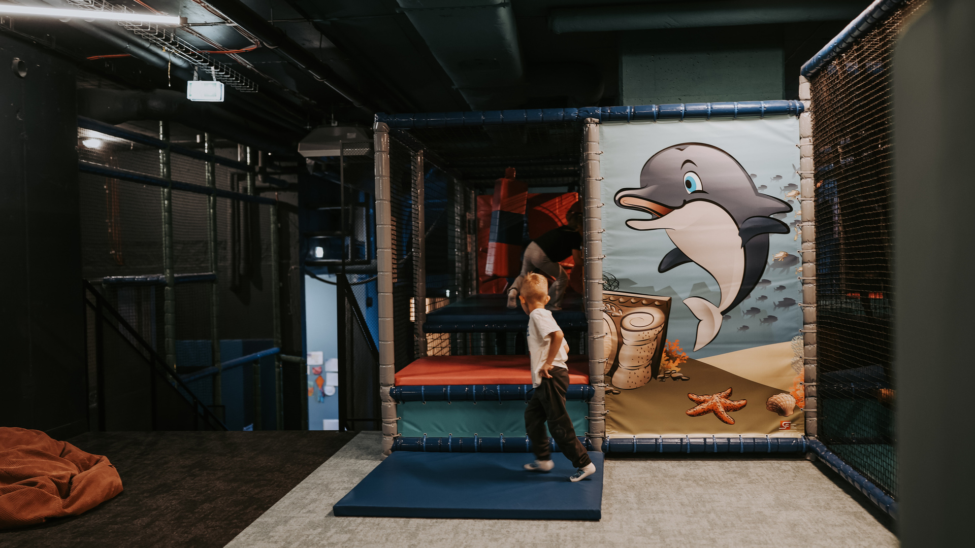 Indoor kids soft-play area with netted climbing frames, ocean mural with dolphin, and children climbing and playing.