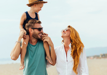 Rodzina spaceruje po plaży w słońcu — dziecko na ramionach ojca, rodzice się śmieją, piasek i wybrzeże w oddali.