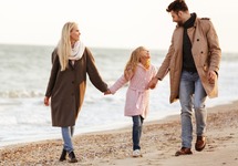 Familie hält Händchen auf kiesigem Strand, Eltern und Tochter in Mänteln bei einem entspannten Küstenspaziergang