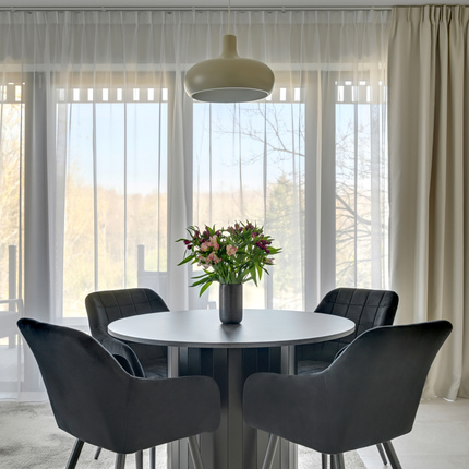 Bright dining nook with round table, vase of fresh flowers, four dark upholstered chairs, pendant lamp and sheer curtains overlooking treetops