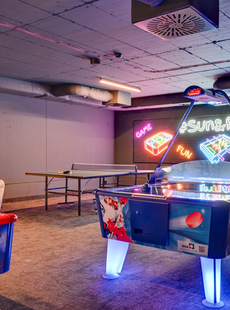 Vibrant hotel game room with illuminated air hockey table, ping-pong and foosball, neon '#Sun&Fun' wall and casual seating