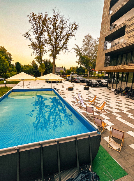Nadziemny basen z leżakami i parasolami na tarasie obok nowoczesnej fasady hotelu i drzew