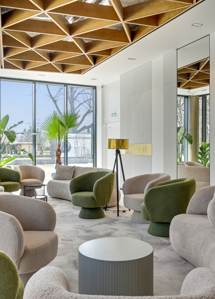 Hotel lounge with plush beige and green chairs, sculpted sofas, floor-to-ceiling windows, gold lamps and geometric wooden ceiling