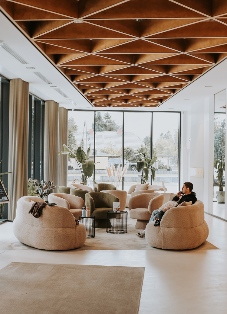 Hotel lobby with plush round armchairs, geometric wooden ceiling, floor-to-ceiling windows, plants and a guest on phone.
