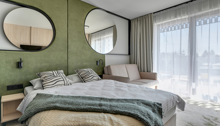 Spacious double room with king bed, green padded headboard with round mirrors, sofa and sunlit balcony