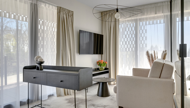 Jasny salon hotelowy z beżowym fotelem, konsolą, telewizorem na ścianie, oknami do podłogi z przezroczystymi zasłonami i bukietem