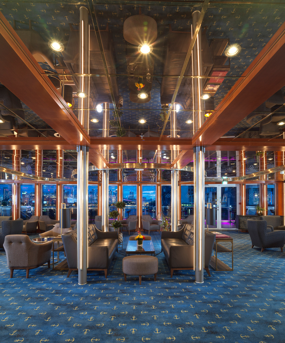 Upscale hotel lounge with mirrored ceiling, warm wood beams, leather sofas, club chairs, blue anchor-pattern carpet and large windows with city views.