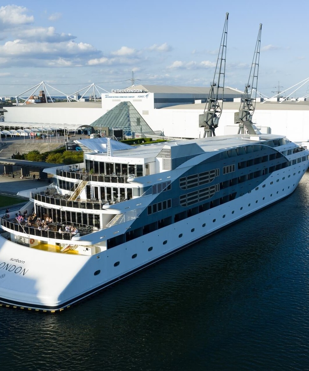Luxury Sunborn London floating hotel docked at ExCeL London, multi-deck terraces with guests dining and waterfront views.