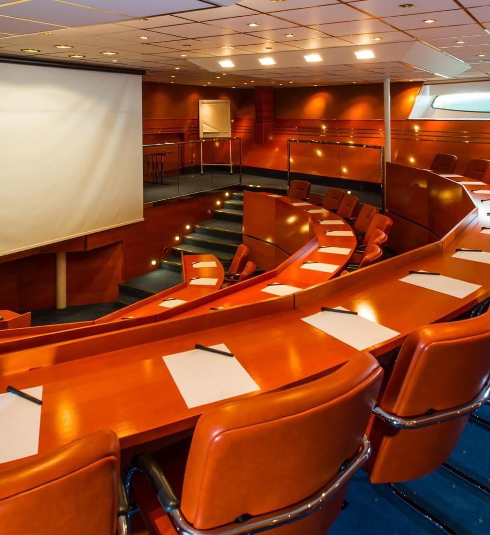 Tiered conference theater with large projection screen, curved wood desks, leather swivel chairs, pens and pads, soft lighting and narrow windows.