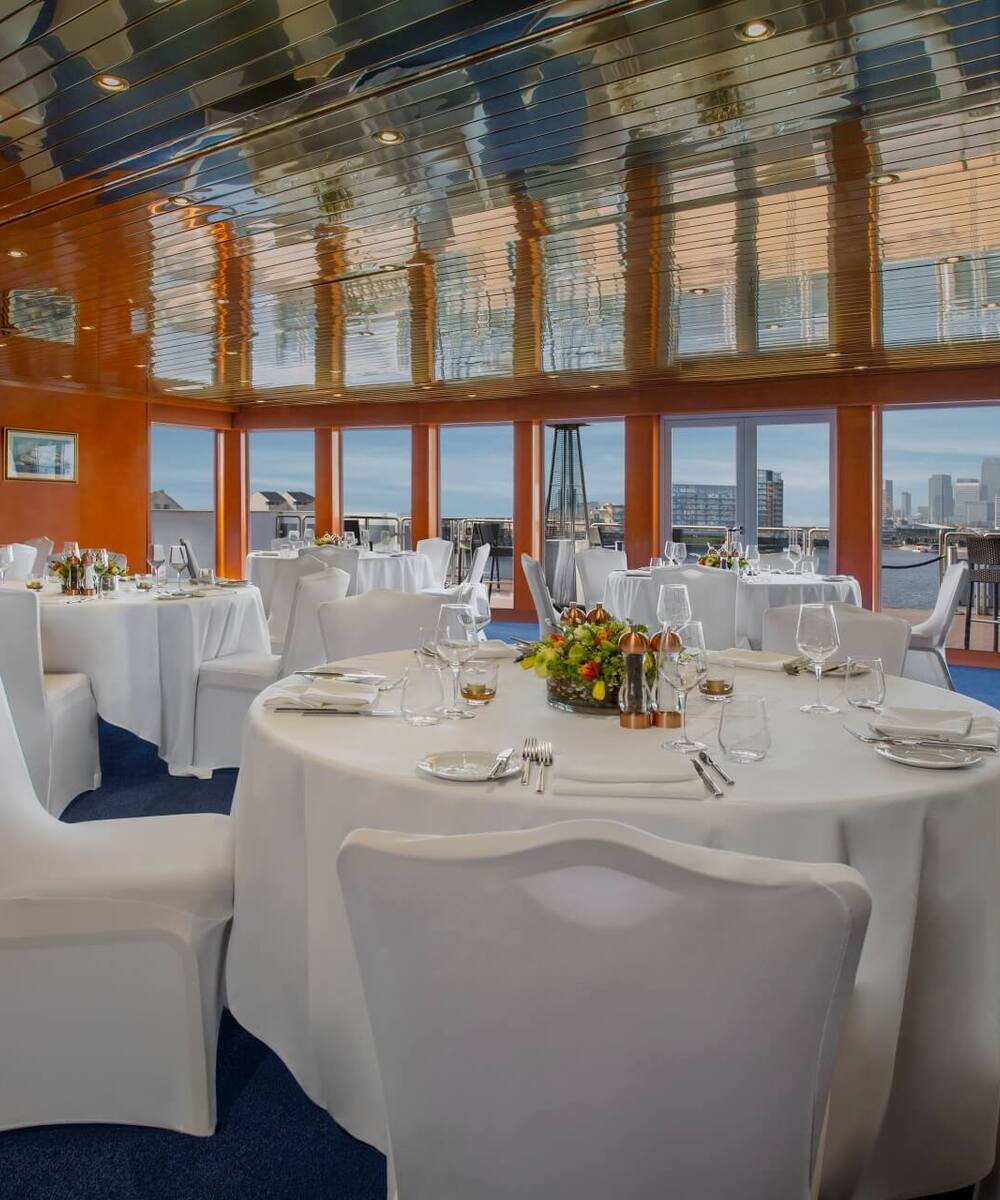 Elegant waterfront dining room with round tables in white linens, floral centerpieces, reflective ceiling and city skyline view