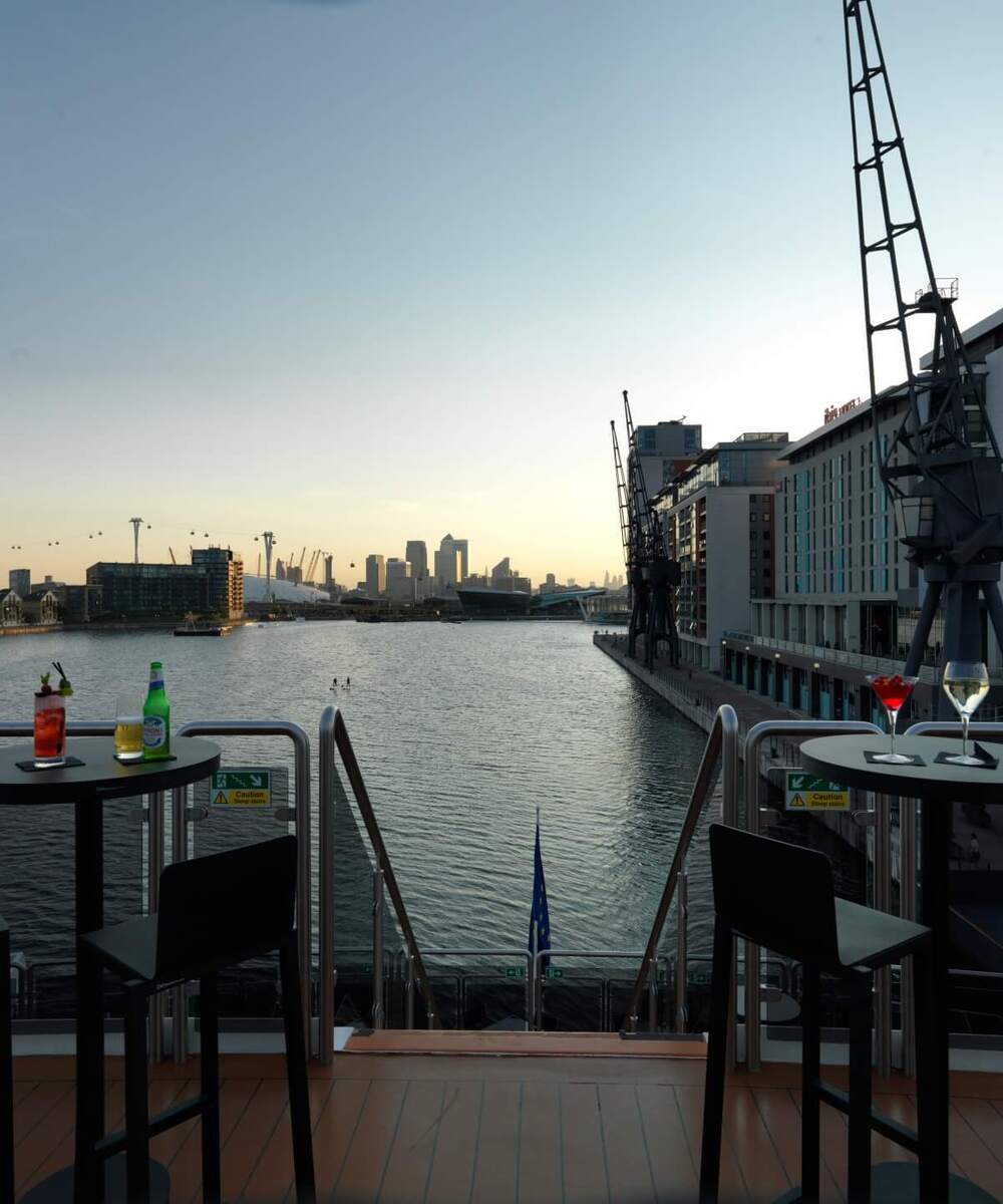 Terrace bar at dusk with cocktails on tables, riverside cranes, Ibis Hotel façade and distant city skyline over calm water