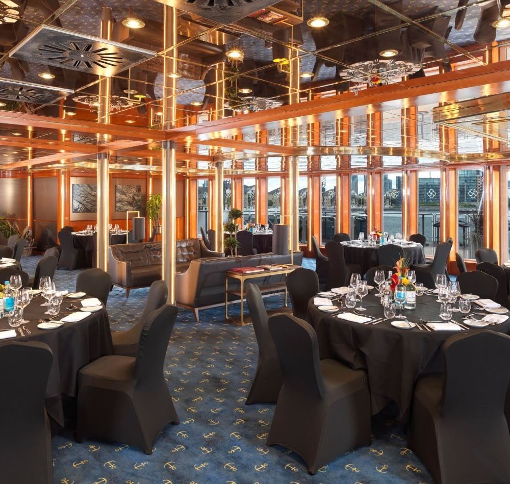 Elegant waterfront banquet room with round black-clothed tables, formal place settings, mirrored ceiling and city skyline views.